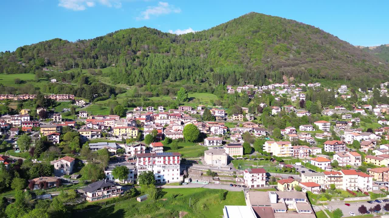 A drone glides over San Fedele near the city of Como, revealing traditional rooftops, narrow streets, green hills, and towering mountains under the summer sun