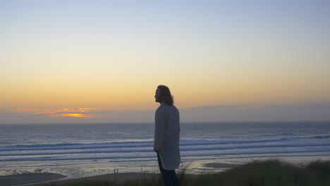 man in kaftan stands on beach watching ocean sunset, wide