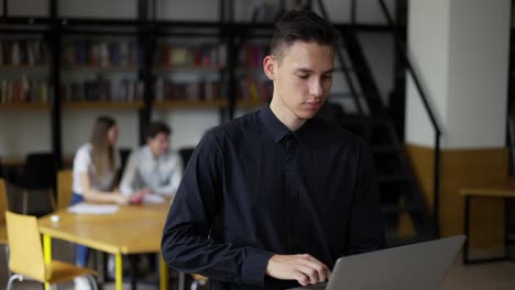 Un-Joven-Estudiante-Serio-Camina-Por-La-Biblioteca-Con-Una-Computadora-Portátil,-Pensando-En-Inspiración,-Encontrando-Soluciones-A-Problemas-E-Ideas.