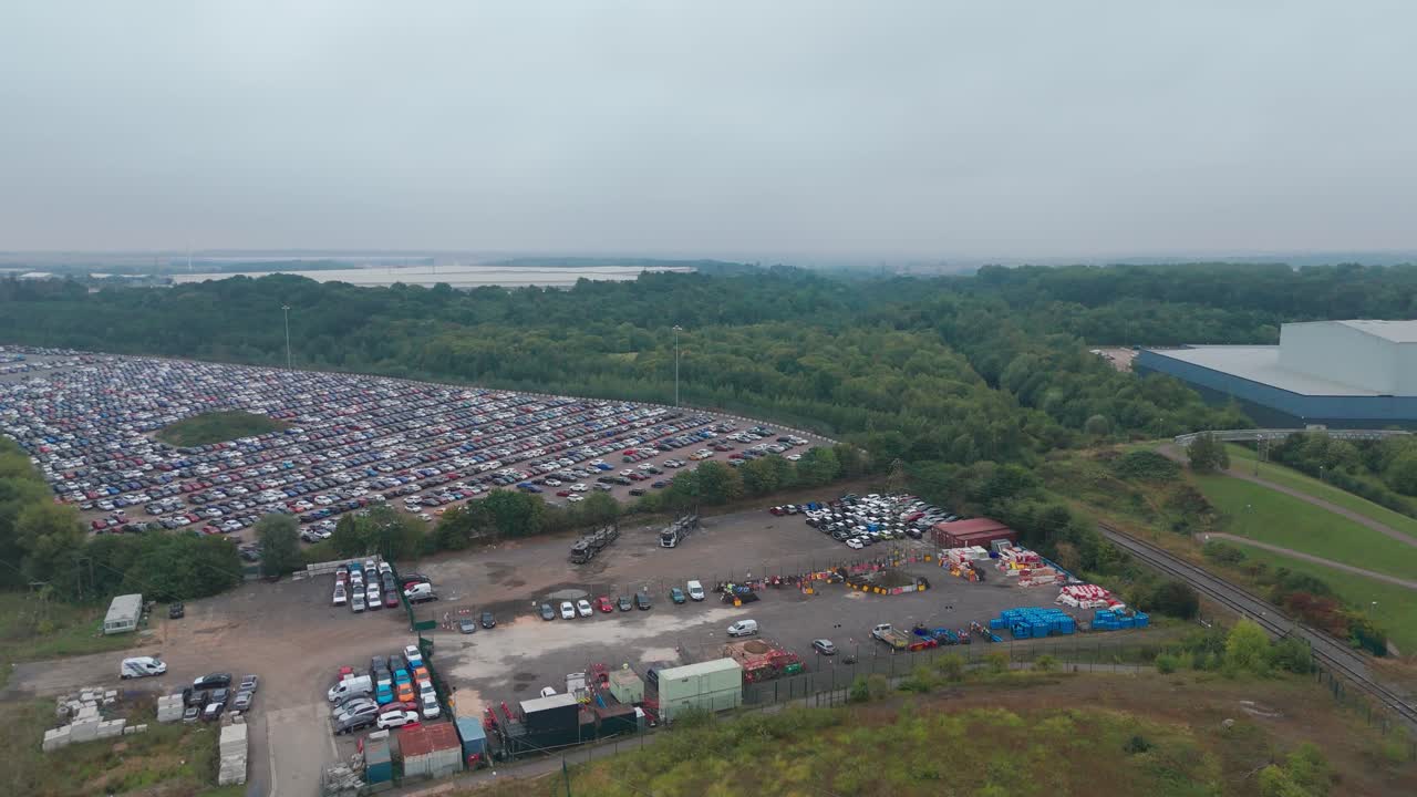 Aerial view of a large parking lot with numerous vehicles and colourful booths, surrounded by greenery and industrial buildings in the distance, indicating an outdoor event or festival
