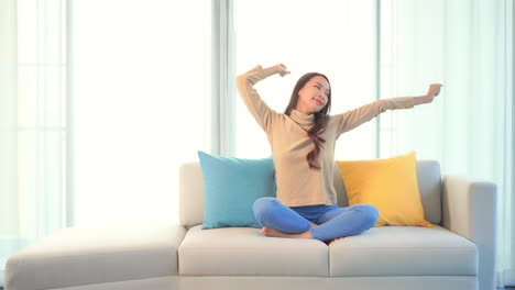 asian young woman stretching her arms over the head while sitting on a sofa having her legs crossed wearing casual clothes daytime -static shot