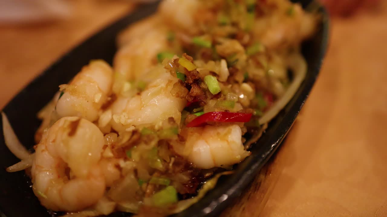 A shallow focus shot of a sizzling prawn and vegetable stir-fry on a black plate, under warm ambient lighting in an Asian restaurant setting