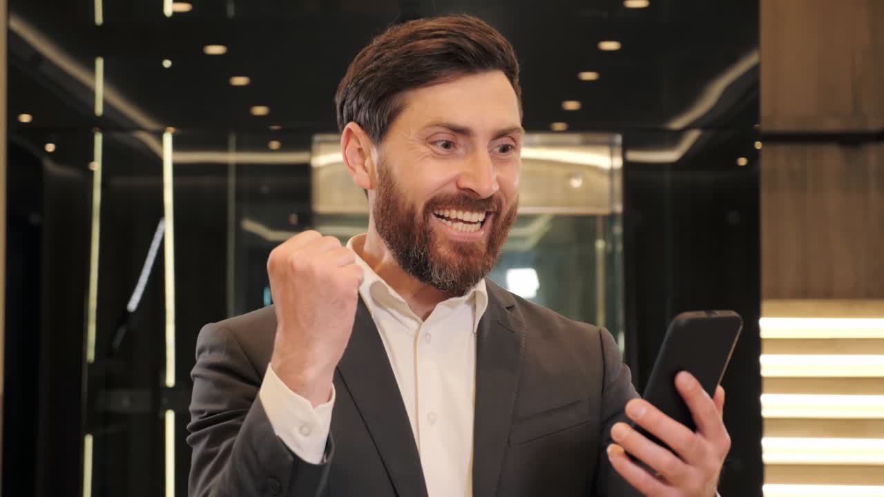 hombre de negocios feliz celebra la victoria mirando en el teléfono inteligente, disfrutando del premio mayor en aplicaciones móviles. hombre de negocios leyendo buenas noticias en el teléfono móvil en el salón moderno. hombre celebrando la victoria.