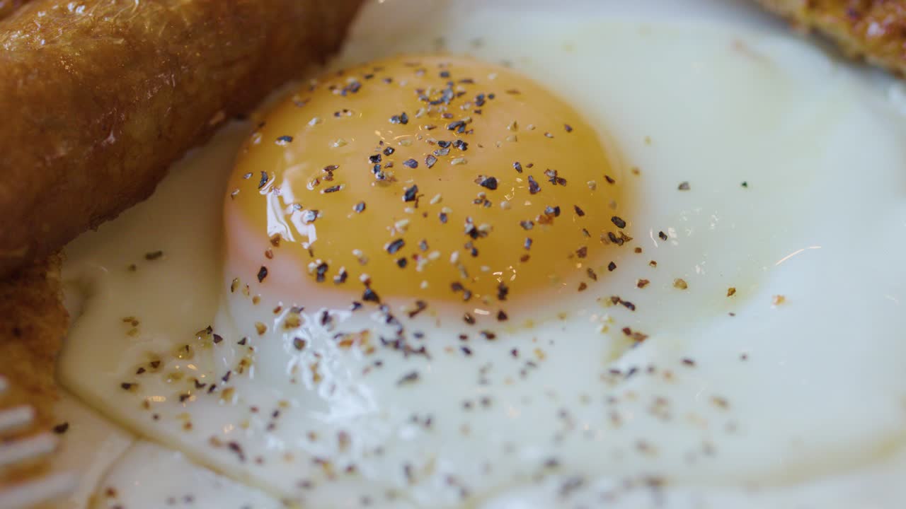 Close-up of fork lifting bacon beside fried egg and sausage, natural lighting, shallow focus
