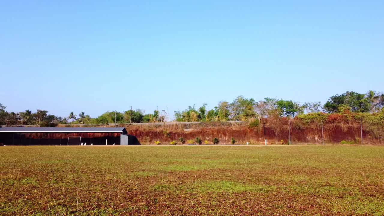 Cinematic pan of a wide green field and rural landscape under a bright blue sky, featuring a rustic shelter and natural earth embankment in a peaceful, sunny outdoor setting