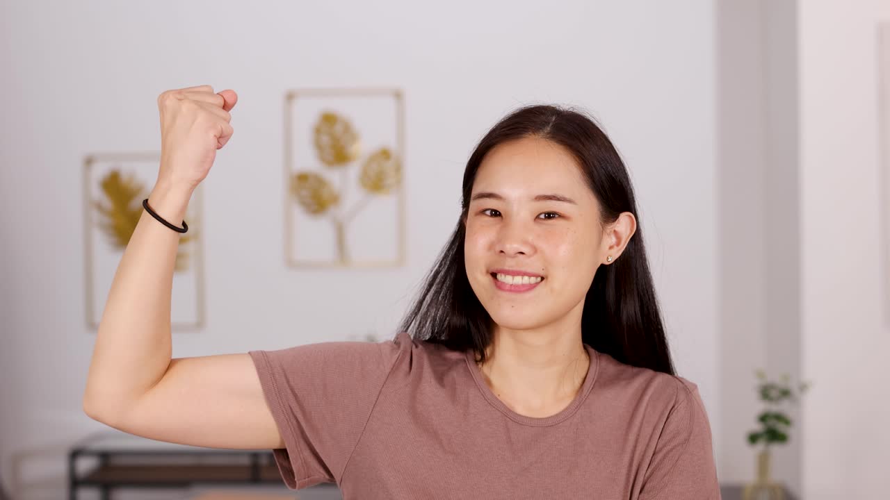 Asian woman smiles and flexes arm in bright home, expressing confidence and positive energy