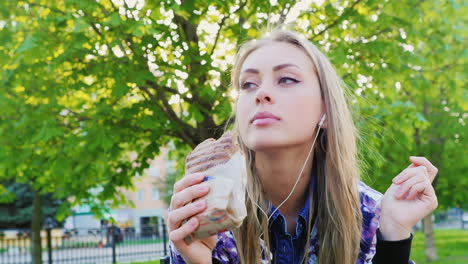 mujer que no come sano comiendo una hamburguesa en el parque escuchando música con auriculares