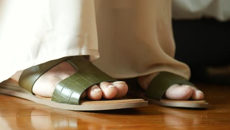 woman's feet in light beige pants wearing olive green sandals on a wooden floor