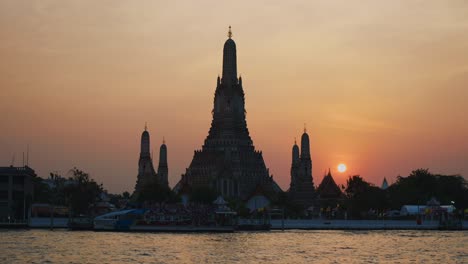 view of wat arun temple in bangkok during a stunning sunset, with a traditional ferry crossing the chao phraya river in front of it