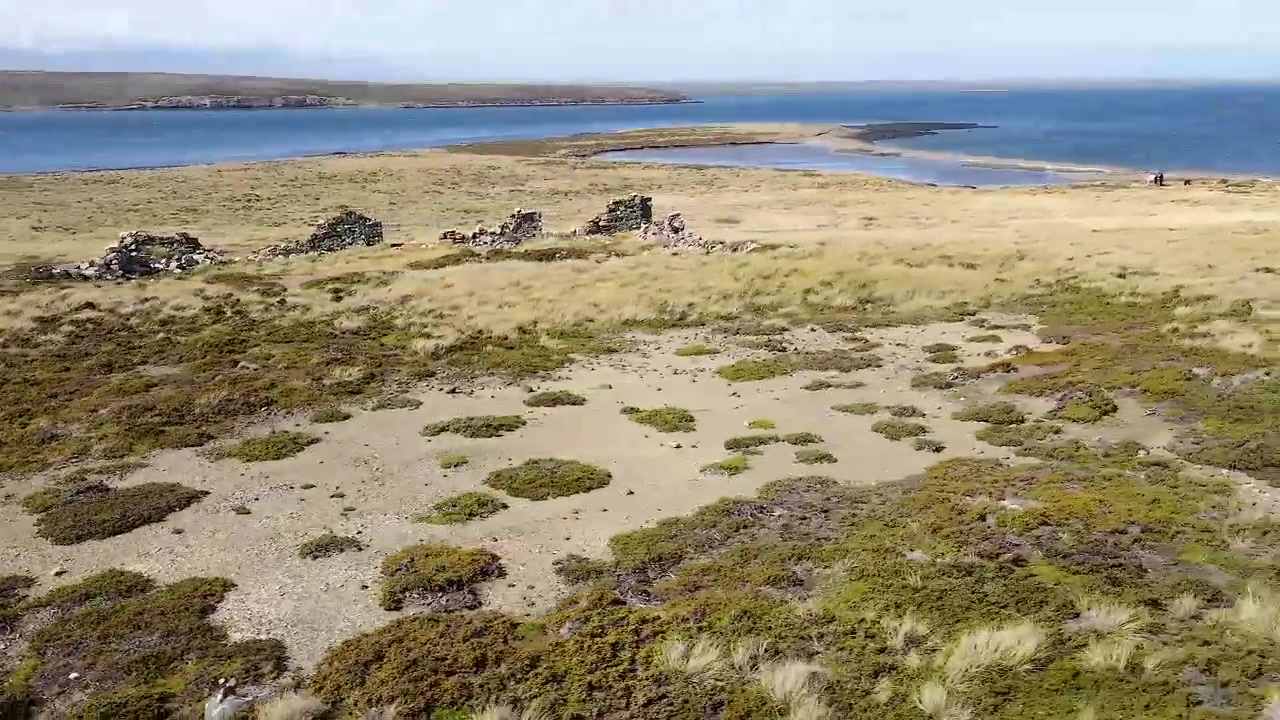 A câmera percorre uma costa relvada com ruínas de pedra, capturando a vastidão da paisagem e os vestígios de uma era passada.