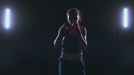 A-beautiful-female-boxer-strikes-directly-into-the-camera-looking-into-the-camera-and-moving-forward-on-a-dark-background-with-a-backlight.-Steadicam-shot