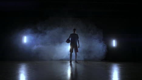 Die-Silhouette-Eines-Basketballspielers-Auf-Dunklem-Hintergrund-Mit-Rauch-Auf-Dem-Basketballplatz-Wirft-Einen-Basketball-Und-Blickt-In-Zeitlupe-In-Die-Kamera