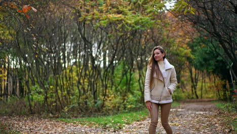 smiling positive lady in white jacket and tight pants walks backwards looking into camera. woman turns around and waves her hair and walks away by the road in autumn park.
