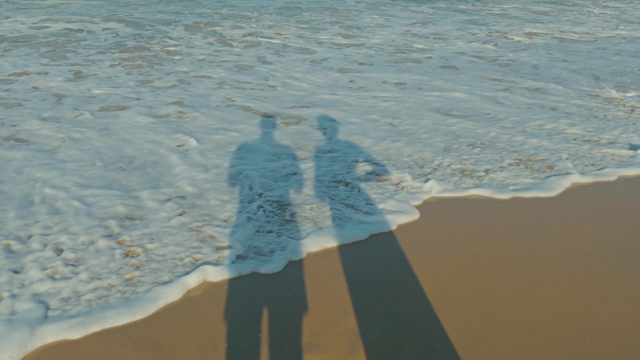 Shadow of a couple on sandy beach as calm waves roll over and recede, creating a romantic and peaceful coastal scene with soft motion and natural light.