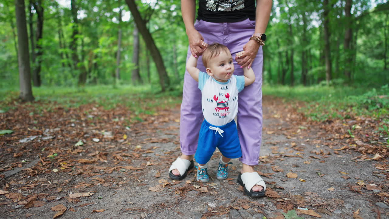 Little toddler stepping slowly looking around. Mommy's holding her baby helping him to walk. Nature blurred backdrop.