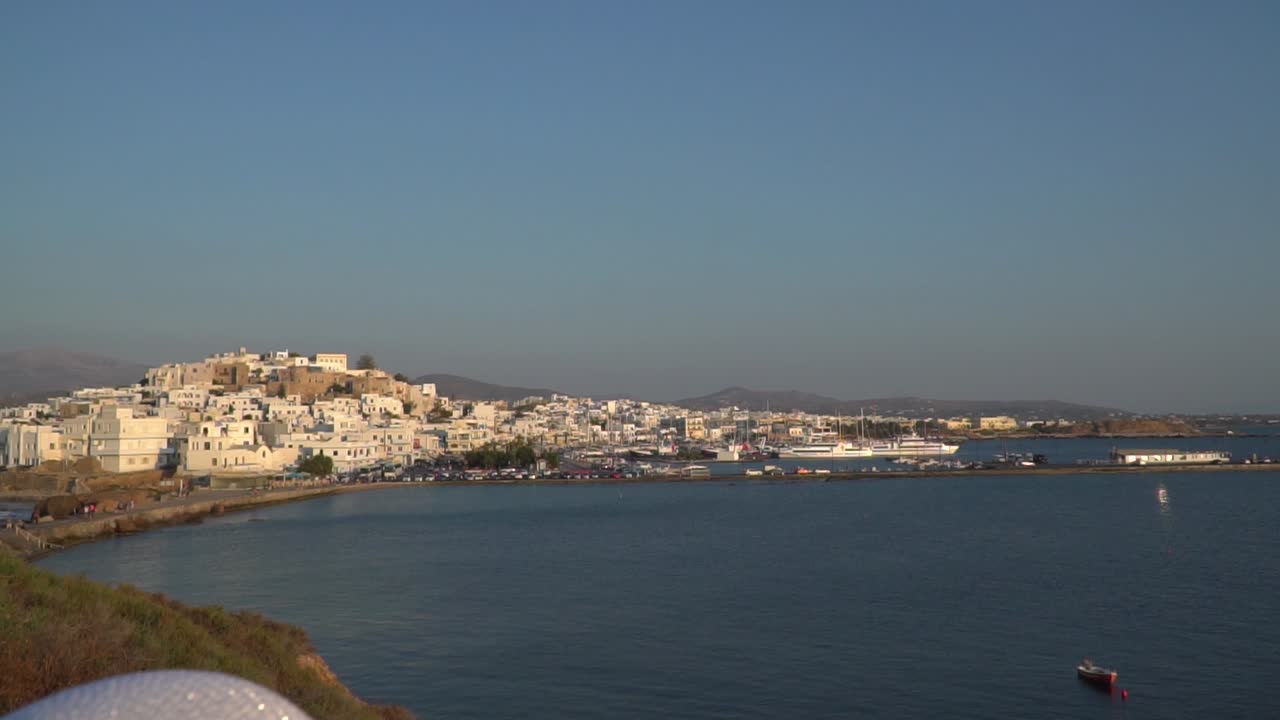 여름 드레스와 거리를 바라보는 모자를 쓴 소녀 뒤에 있는 낙소스 마을의 모습을 위로 기울이세요