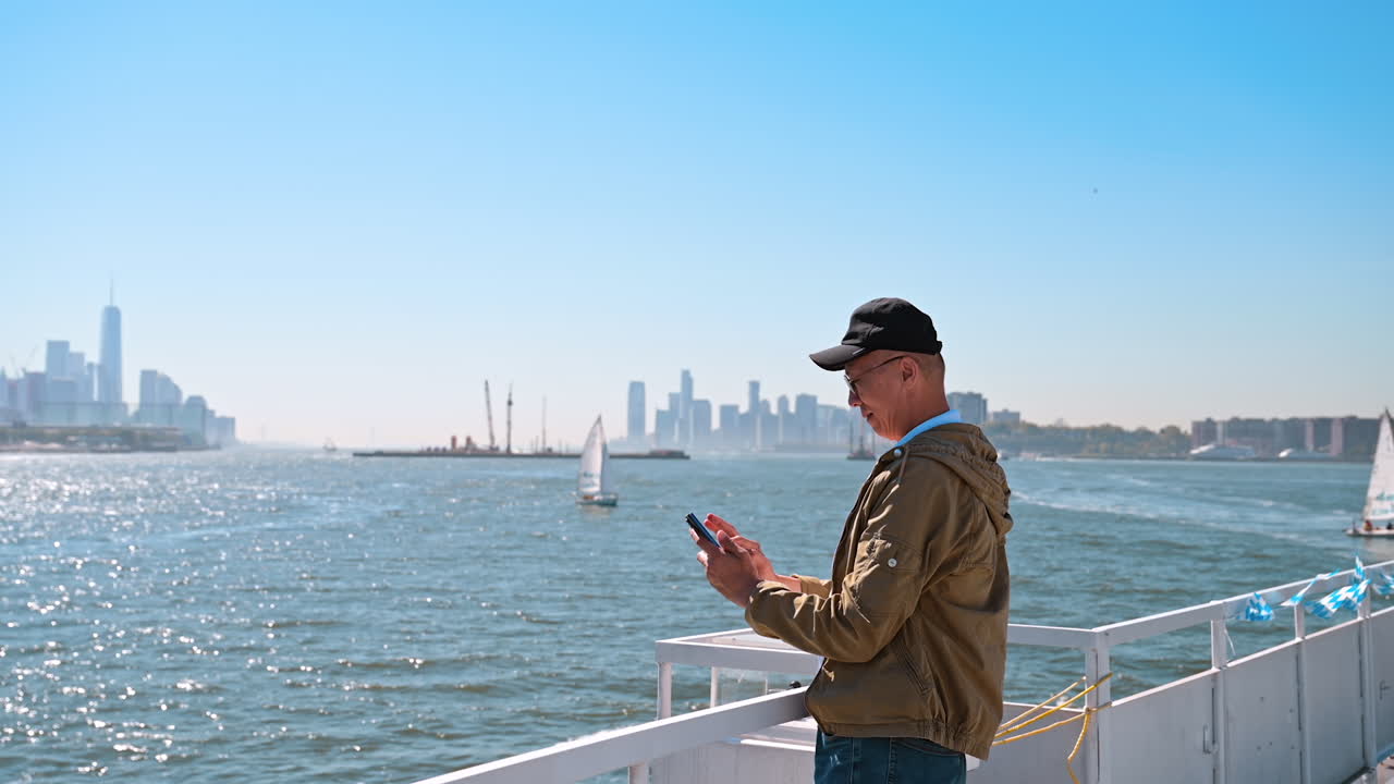 New York, USA, 9 August 2025: Man using smartphone on ferry with Manhattan skyline view. A man stands on a ferry looking at his smartphone with the skyline of Manhattan and sailboats in the background