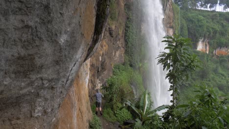 Toma-En-Cámara-Lenta-De-Un-Excursionista-Caminando-Por-Un-Pequeño-Sendero-Del-Acantilado-Debajo-De-Una-Cascada-Tropical