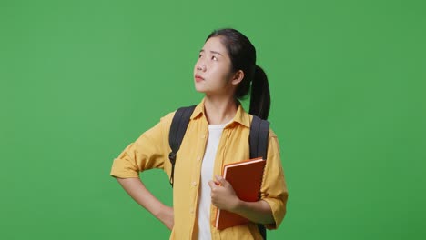 asian woman student with a backpack and a book standing with one arm akimbo and looking around in the green screen background studio