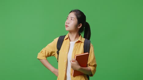 백과 책을 들고 한 팔을 고 서 있는 아시아 여성 학생이 녹색 스크린 배경 스튜디오를 둘러보고 있다.