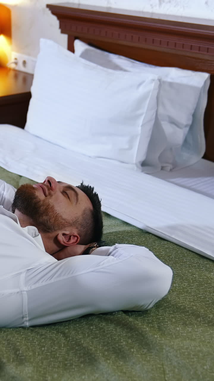 Businessman rest at daytime. Handsome man in white shirt and black trousers lying on bed in comfortable hotel. Adult male relaxing during business trip. Vertical video