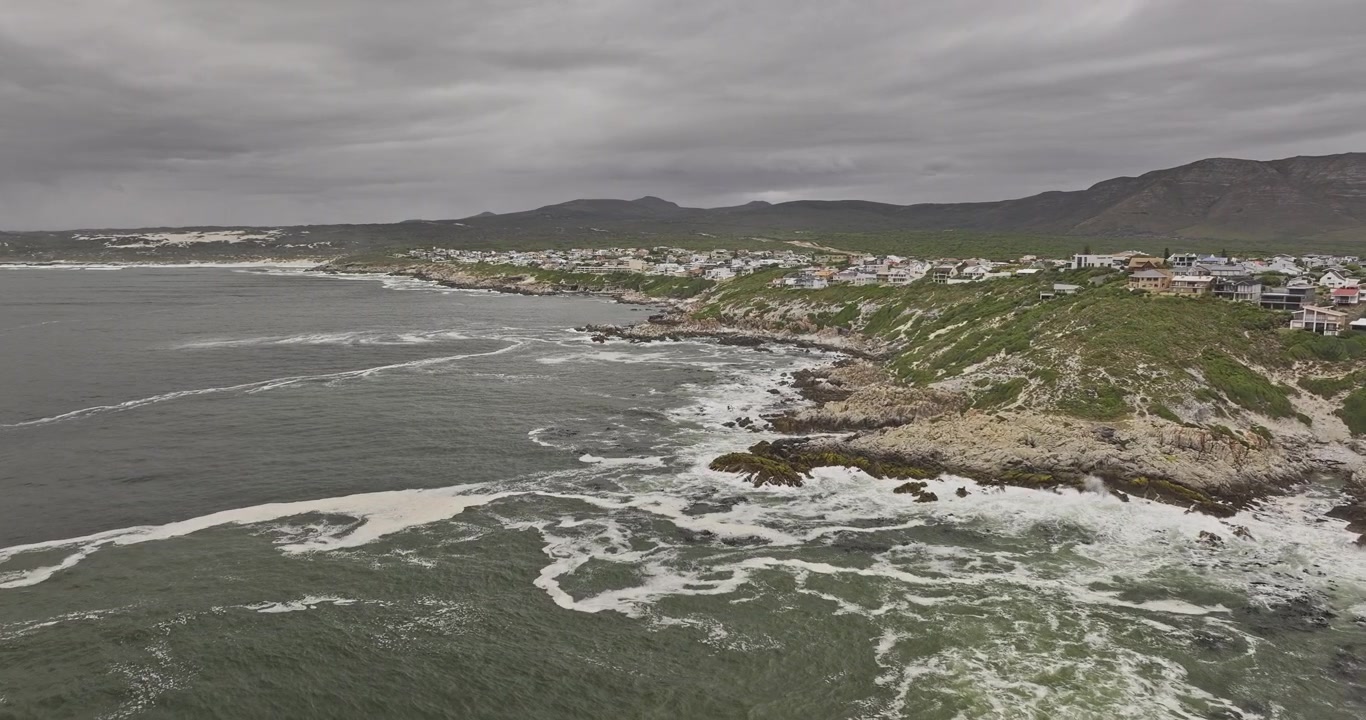 Gansbaai, Sudáfrica. Sobrevuelo aéreo bajo v2 alrededor de las casas de De Kelders sobre una costa rocosa con olas, montañas y cielos nublados - Filmado con Mavic 3 Pro Cine - 7 de enero de 2024