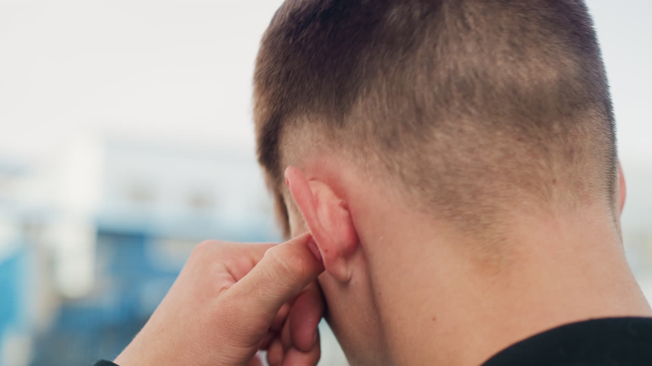 Primer plano de un hombre joven insertándose un tapón para los oídos. Manos ajustando el tapón de espuma en la oreja, pelo corto, fondo urbano difuminado, preparación rutinaria para entornos ruidosos, contextos de trabajador, viajero o músico, seguridad