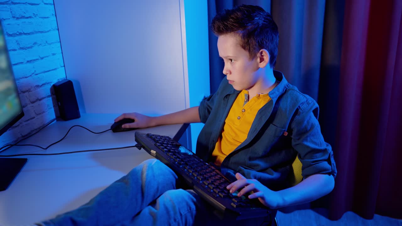 Children's entertainment. Boy is playing video games at home. Teenage boy sitting at the desk with a keyboard on legs and concentrated on online games on his computer.