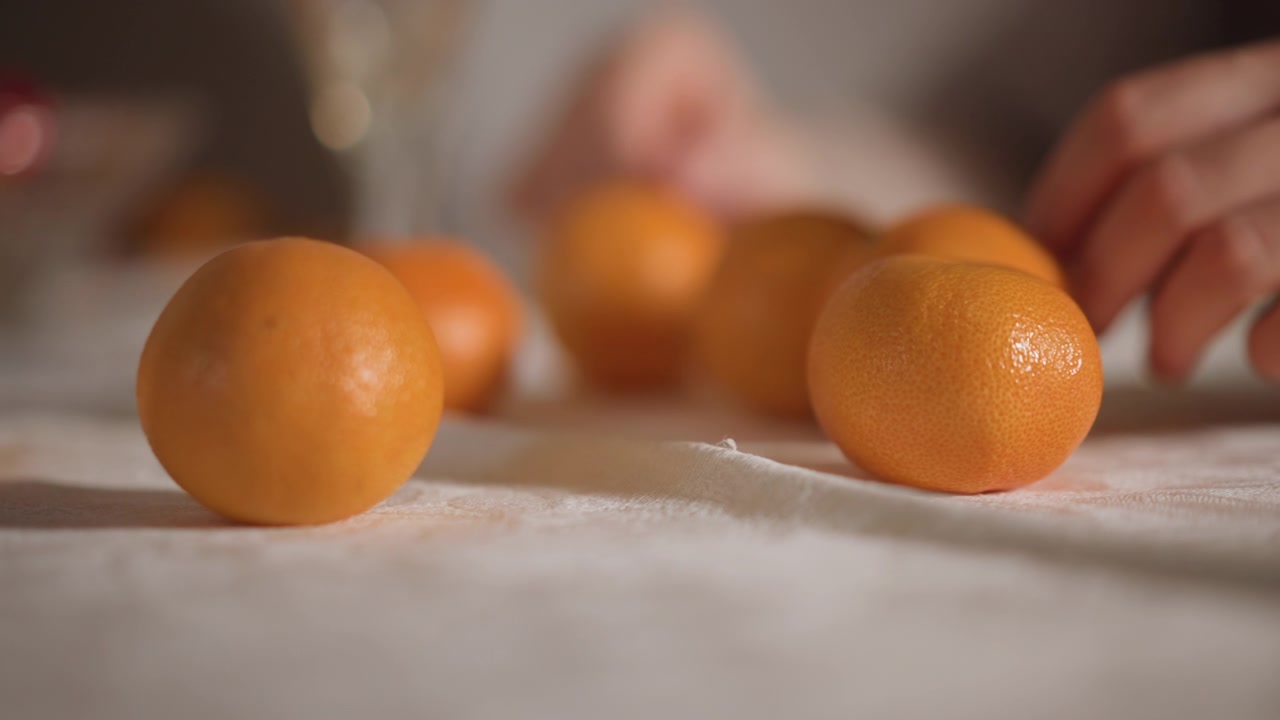 Close-up de tangerina em mesa de linho, luz natural suave, profundidade de campo rasa, mão alcançando ao fundo, várias tangerinas rolando suavemente sobre tecido texturizado, tons quentes e suaves, ambiente doméstico acolhedor.