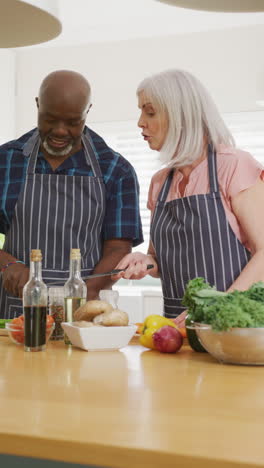 vertical video of happy diverse senior couple preparing food together and talking in kitchen