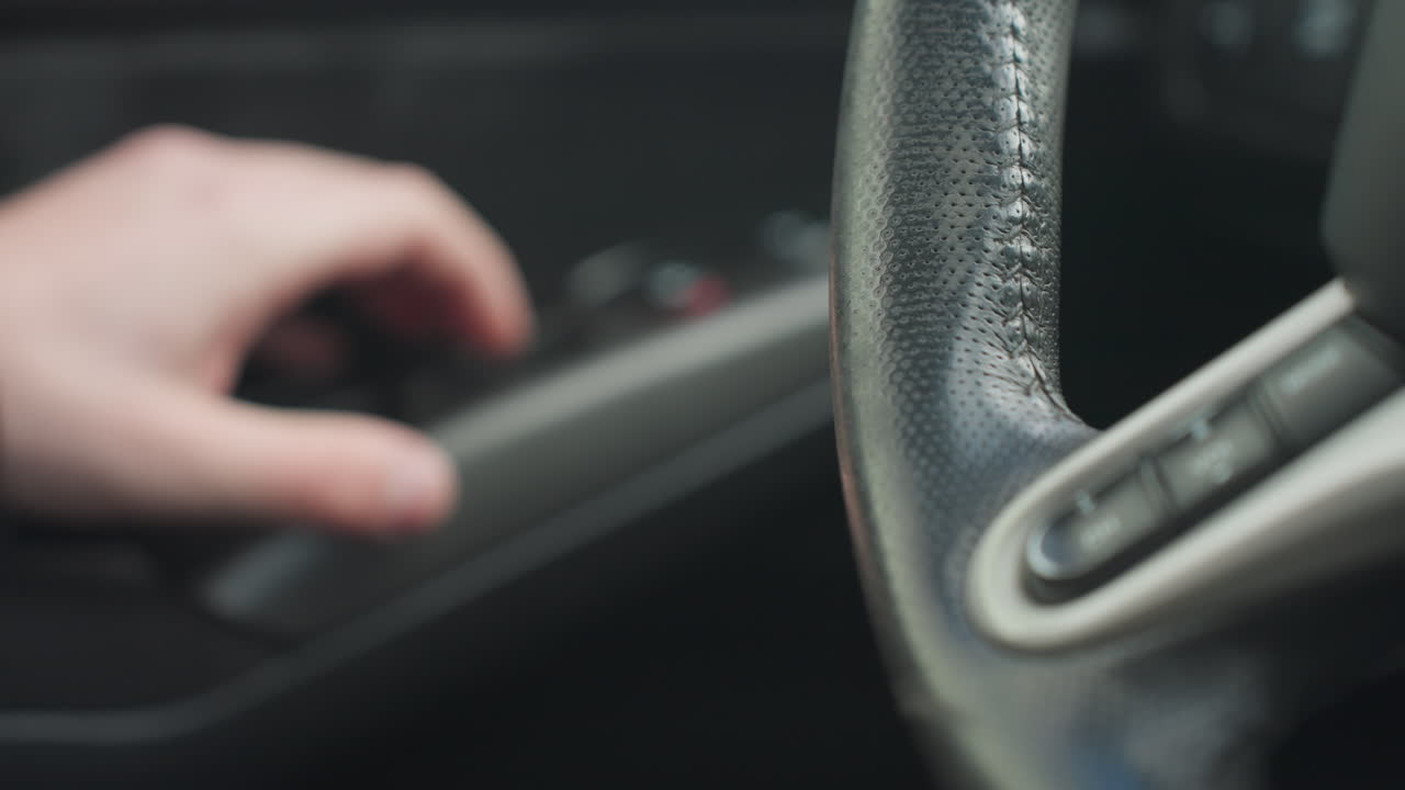 close up of person hand resting beside textured steering wheel while lightly reaching toward door controls inside vehicle, capturing tactile moment and fine interior car detail in soft focus