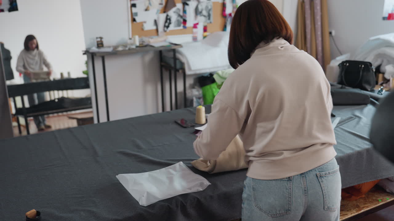 Stylist in bright sewing workspace delicately folds beige cloth removed from mannequin, sliding piece into transparent packaging bag on large cutting table while mirror reflects precise motions