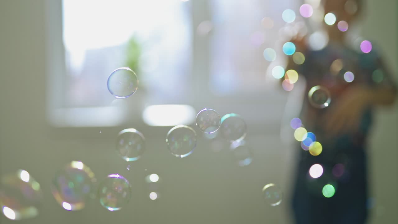 Round colorful bubbles flying in the room on the blurred background with a boy. Beautiful soap bubbles and a silhouette of a boy playing with them indoors.