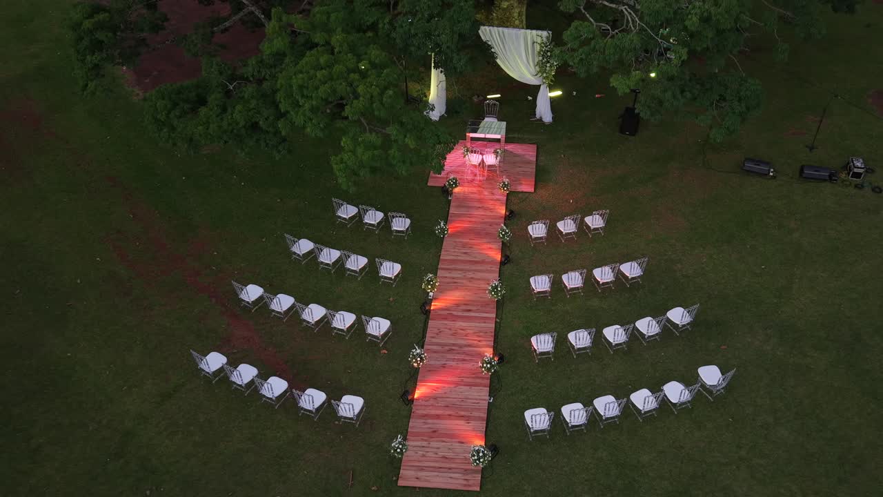 Drone che scende in picchiata sopra un allestimento per cerimonia nuziale all'aperto con passerella in legno, sedie bianche e altare decorato in un contesto di giardino.