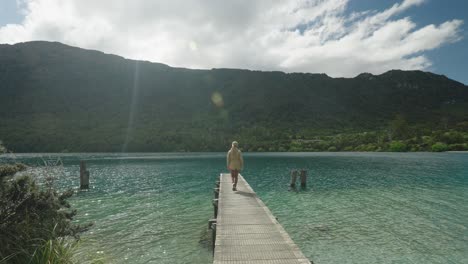 woman walking on wooden jetty at pristine blue cove with mountains, new zealand