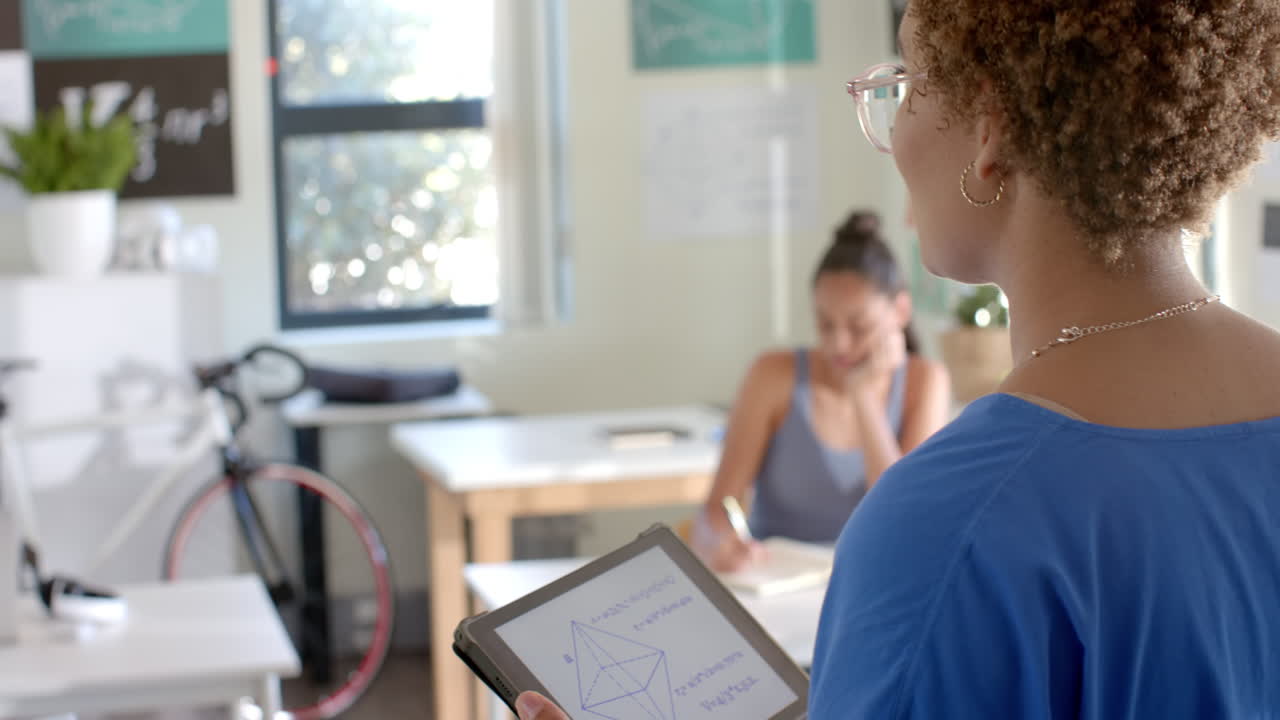 Holding tablet with geometric shapes, teacher guiding student studying in classroom