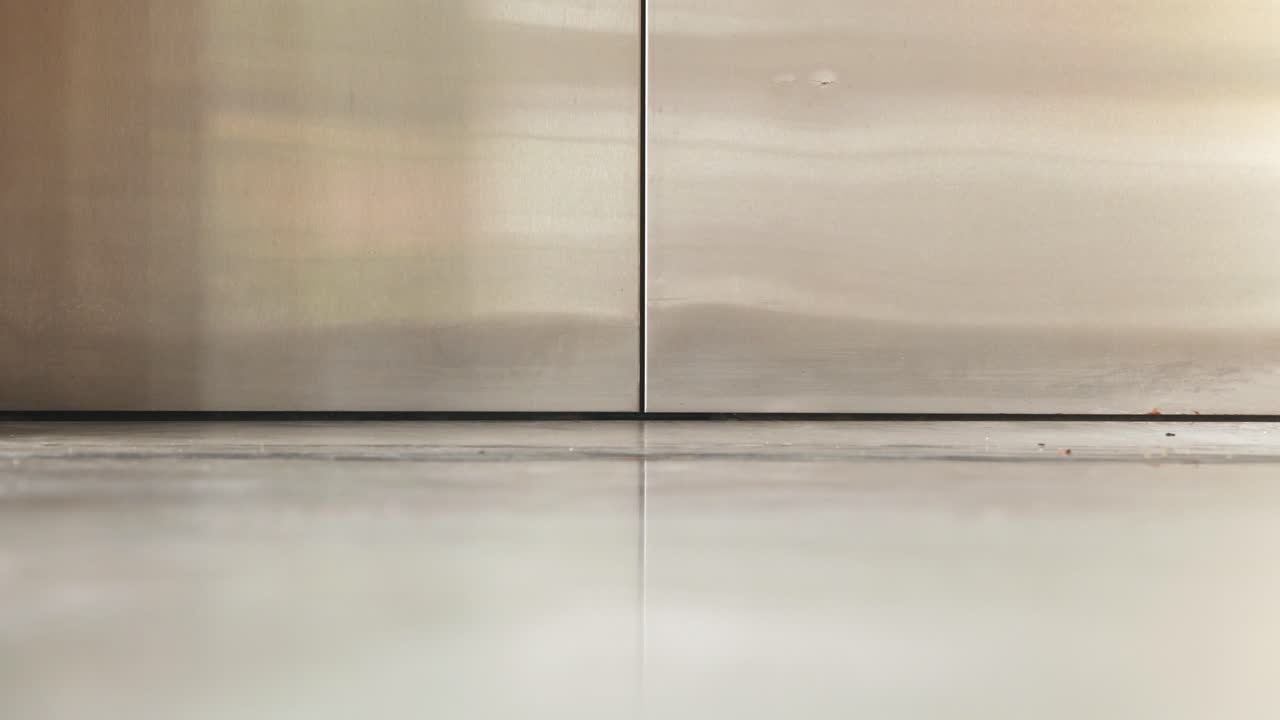 A very low-angle, close-up view of a metal elevator door opening onto a shiny concrete or polished floor, highlighting the gap and threshold