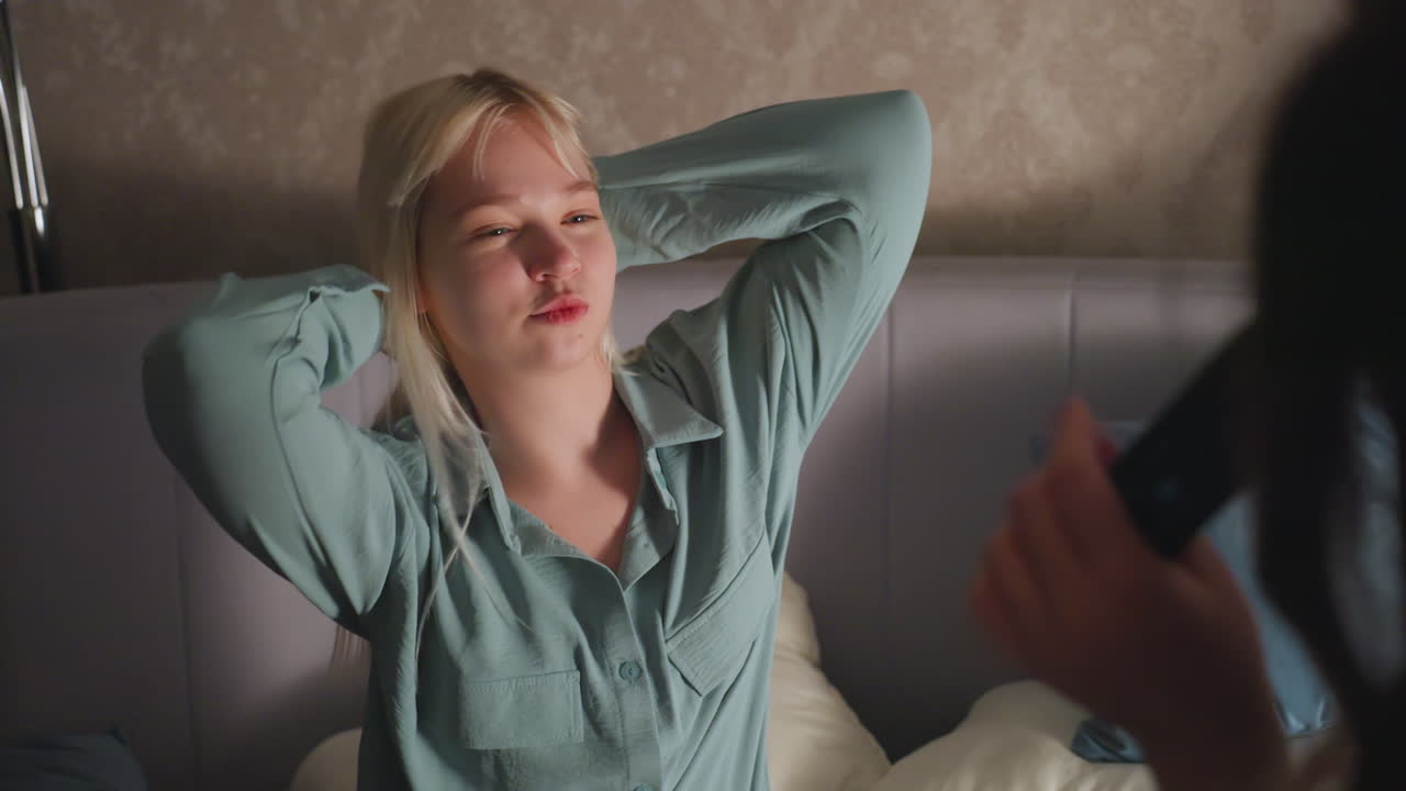 Close up of siblings sitting on bed during playful evening as girl in black pajamas sings using remote while sister watches with smile, enjoying fun bonding moment in cozy bedroom atmosphere