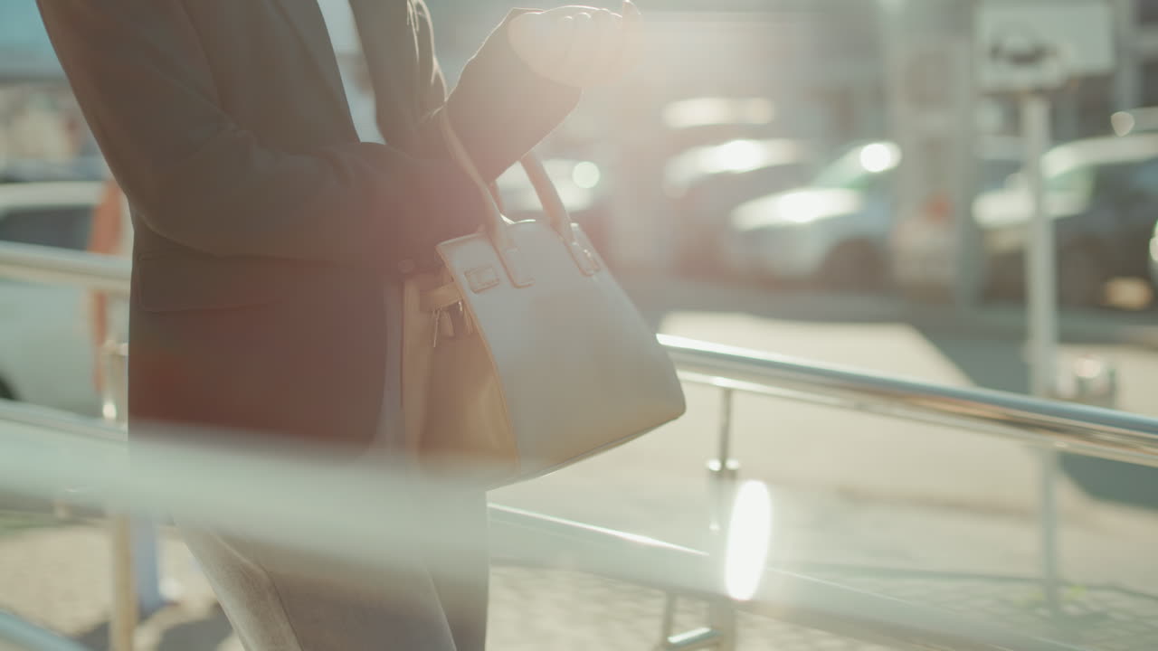 Woman in business outfit retrieving car key from handbag near railing in sunlit urban area, second woman walking in background, parked cars visible