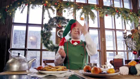 Hungry elf drinking at the table at Christmas. Male elf in green costume with hood eating at festive table on decorated background. New Year and Christmas concept.