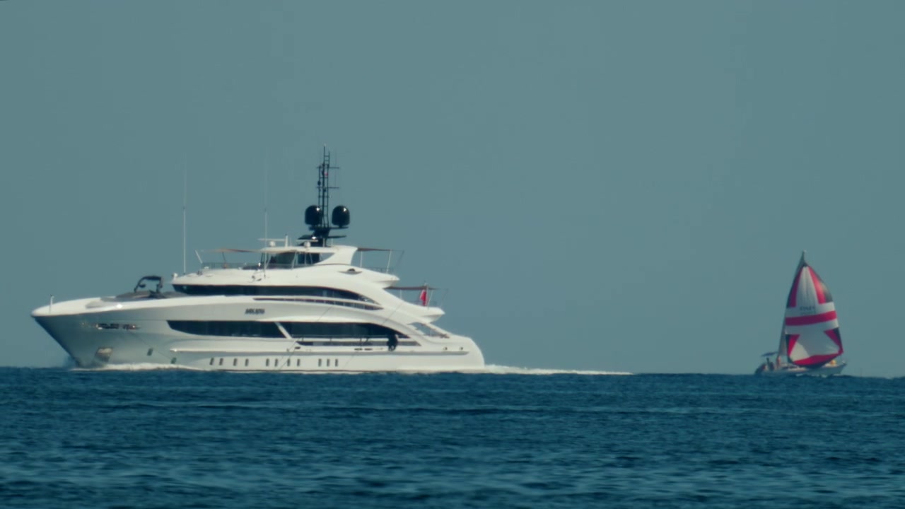 Un grande yacht a motore che naviga velocemente accanto a una barca a vela più piccola su acqua blu calma sotto un cielo estivo limpido, Antibes, Francia