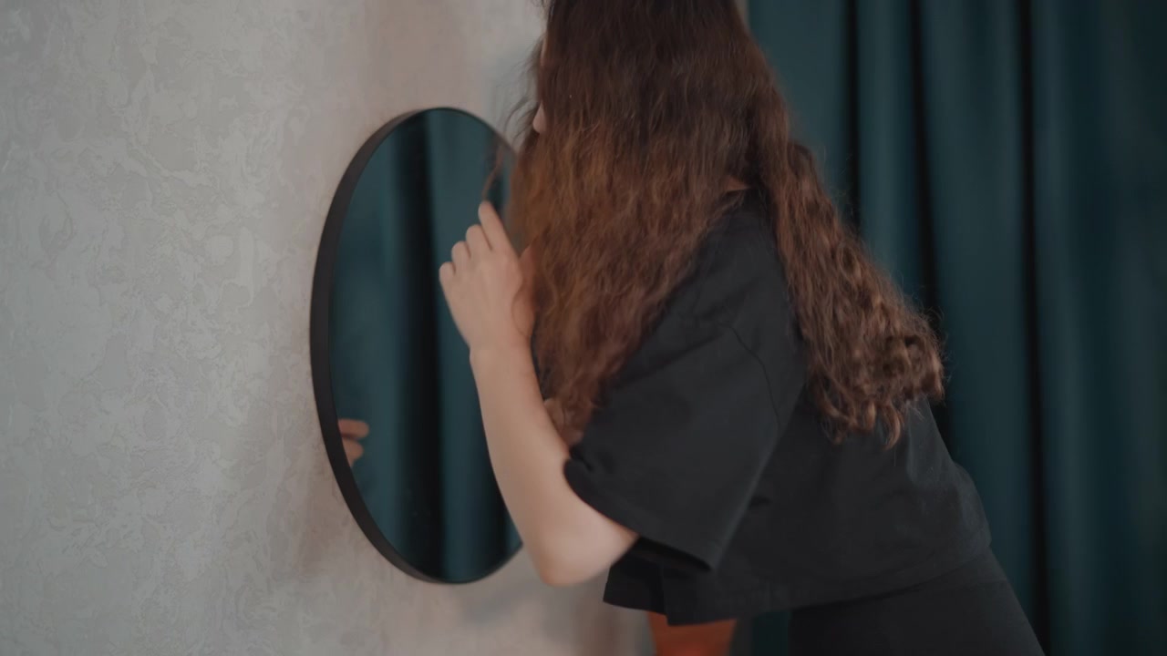 Donna adulta con capelli ricci si china verso uno specchio rotondo a parete, alzando la mano mentre si trucca in una zona trucco illuminata delicatamente, mostrando una cura attenta, igiene personale e una preparazione calma.