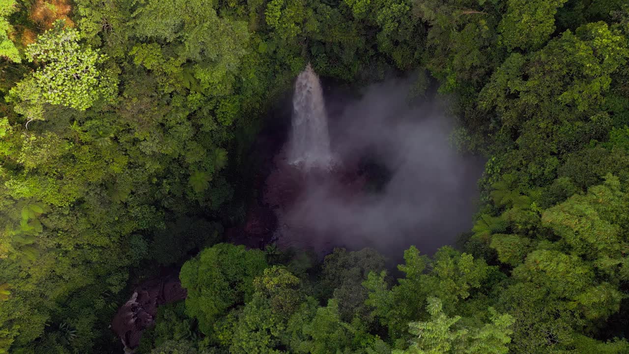 Nungnung Waterfall is hidden deep in the jungle and surrounded by lush green trees with steam rising from the crashing water as seen from an aerial drone perspective in the tropical forest.