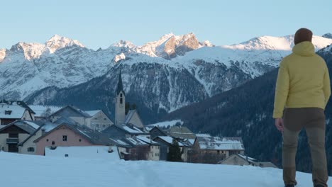 young caucasian man in yellow jacket and beanie walking and stopping to see view of small mountain village and church in guarda, switzerland