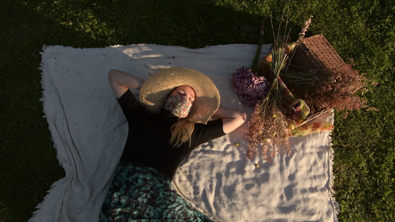 Woman wearing a face mask lying on picnic blanket on the grass waves arms