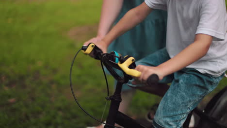un primer plano de las manos de una mujer mientras ayuda a su hijo a montar una bicicleta, en un fondo verde