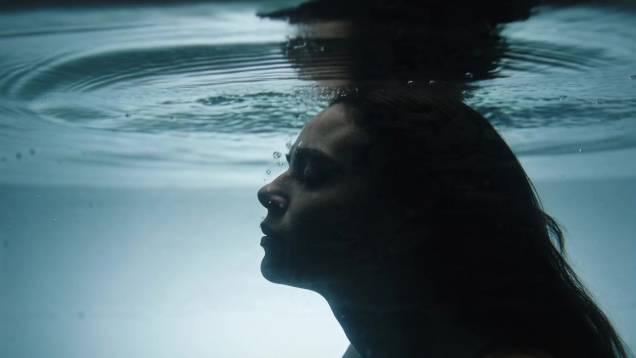 Mulher adulta exalando, de maiô escuro, com a cabeça debaixo da superfície da piscina, formando bolhas e ondulações.