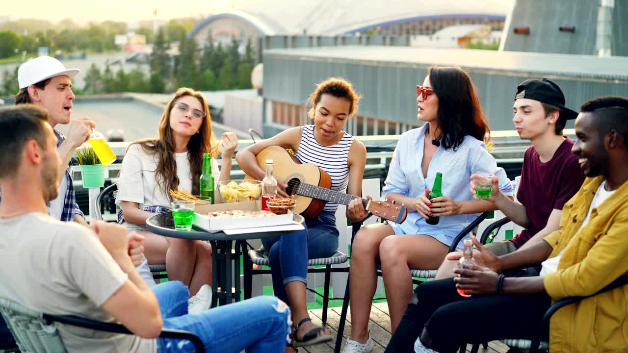Attractive African American girl is playing the guitar, her friends are singing, drinking and eating at table on roof enjoying weekend and warm sunny day.