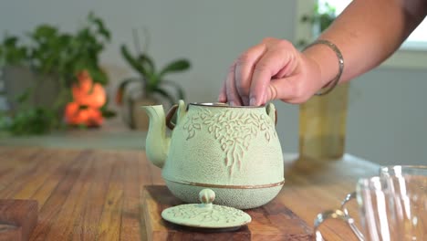 making herbal tea in a green teapot on a wood table in a light and airy room with green plants in the background and a glass teacup