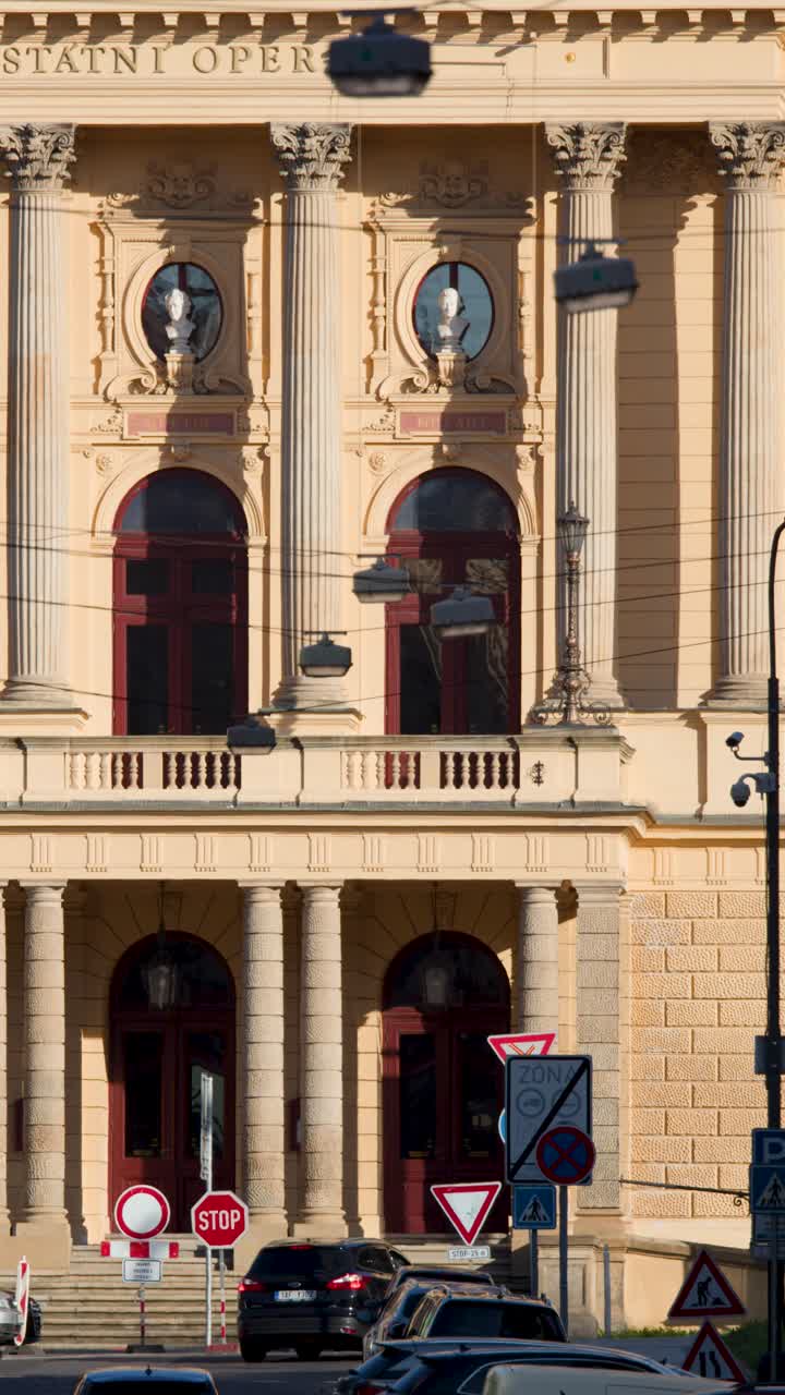 Vehículos pasan por delante de un histórico edificio de ópera neoclásico, la luz del sol resalta detalles arquitectónicos ornamentados, plano fijo